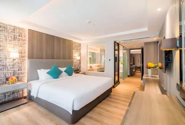 Modern hotel bedroom with a king bed, teal pillows, bedside lamps, wall TV, and a visible bathroom.