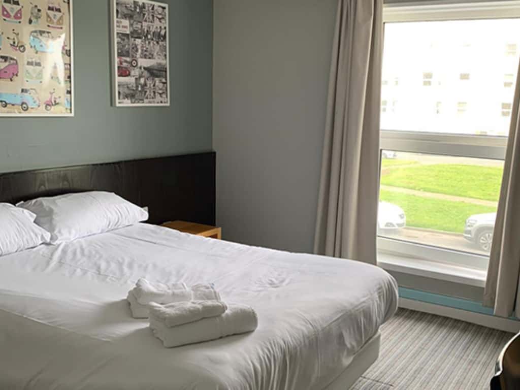 A white bed with folded towels sits next to a window with grey curtains and two framed pictures on a teal wall