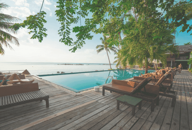 Guests relax on lounge chairs on a wooden deck next to an infinity pool overlooking the ocean.