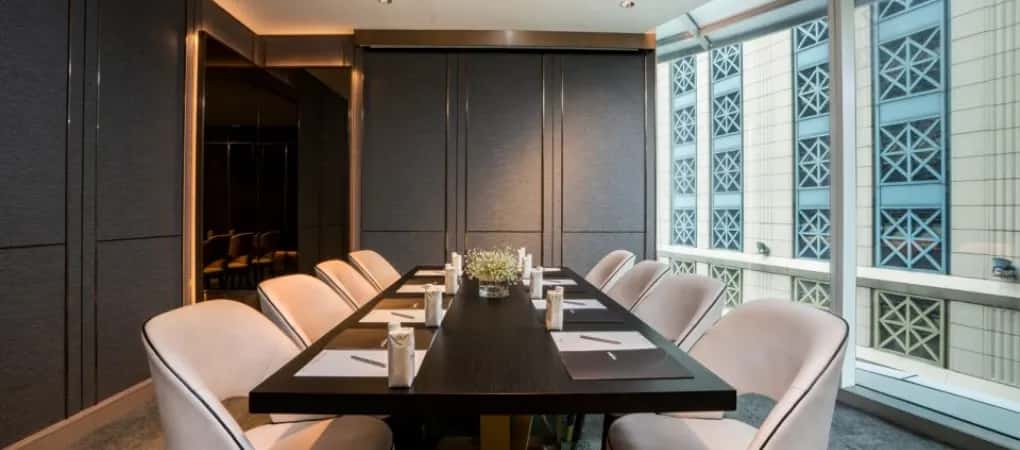 A meeting room with a long dark table, white chairs, place settings, and a window overlooking a building with patterned windows.