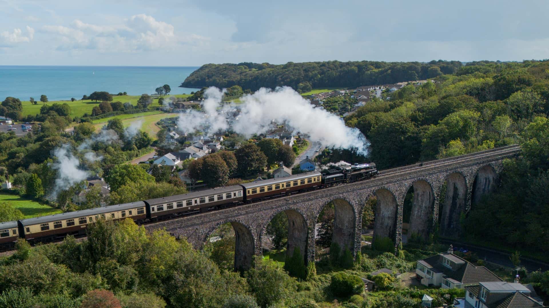 Steam train crossing a tall stone arched viaduct through a green coastal landscape with the ocean in the background