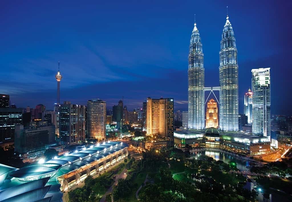 Twin skyscrapers and a broadcast tower illuminate a city skyline at dusk, with a park and convention center.