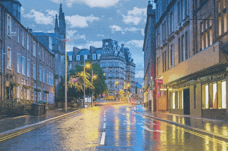 Wet urban street at dusk with historic buildings, streetlights, and reflections on the road.