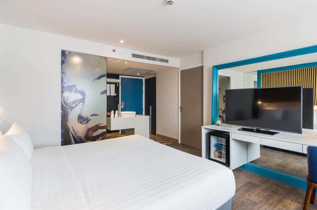 Hotel room with white bed, flat-screen TV, blue-framed mirror, and open bathroom area