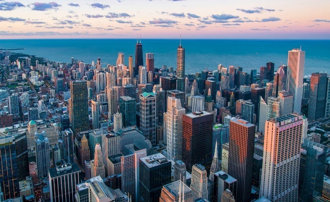 Aerial view of a city skyline with numerous skyscrapers along a large body of water under a cloudy sky.