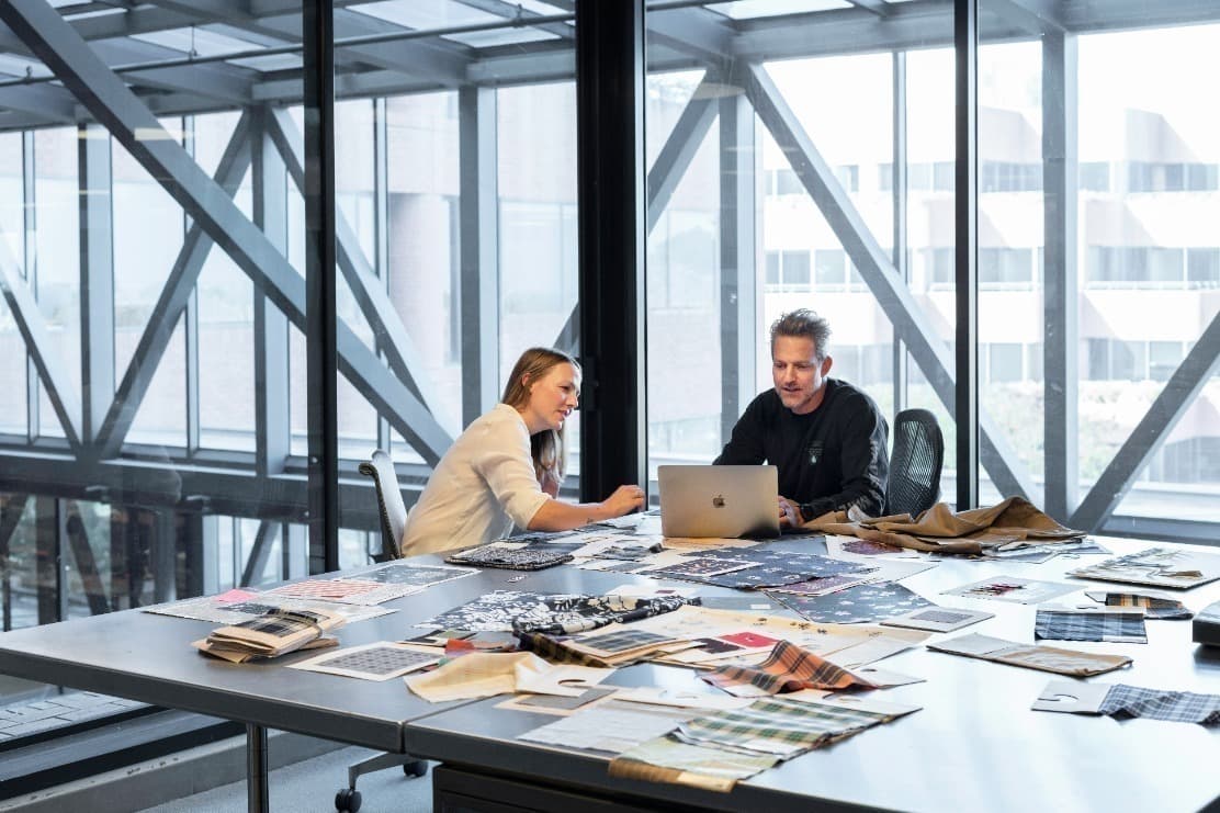 Two people sit at a large table covered in fabric swatches while looking at a laptop in a bright office space