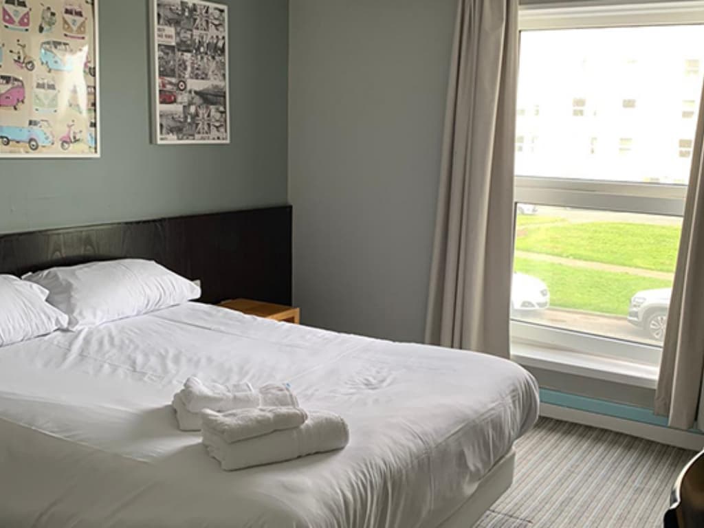 A white bed with folded towels sits next to a window with grey curtains and two framed pictures on a teal wall