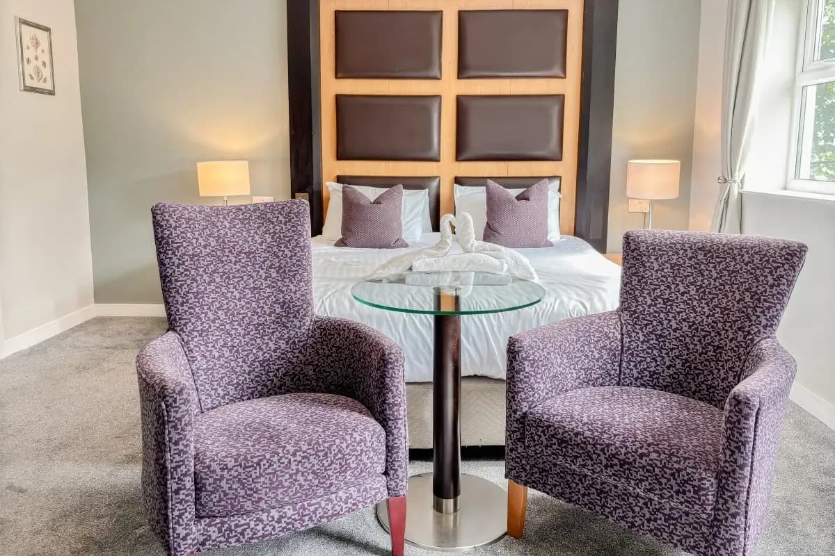 Two patterned purple armchairs sit in front of a bed with a high brown headboard and a glass-topped table