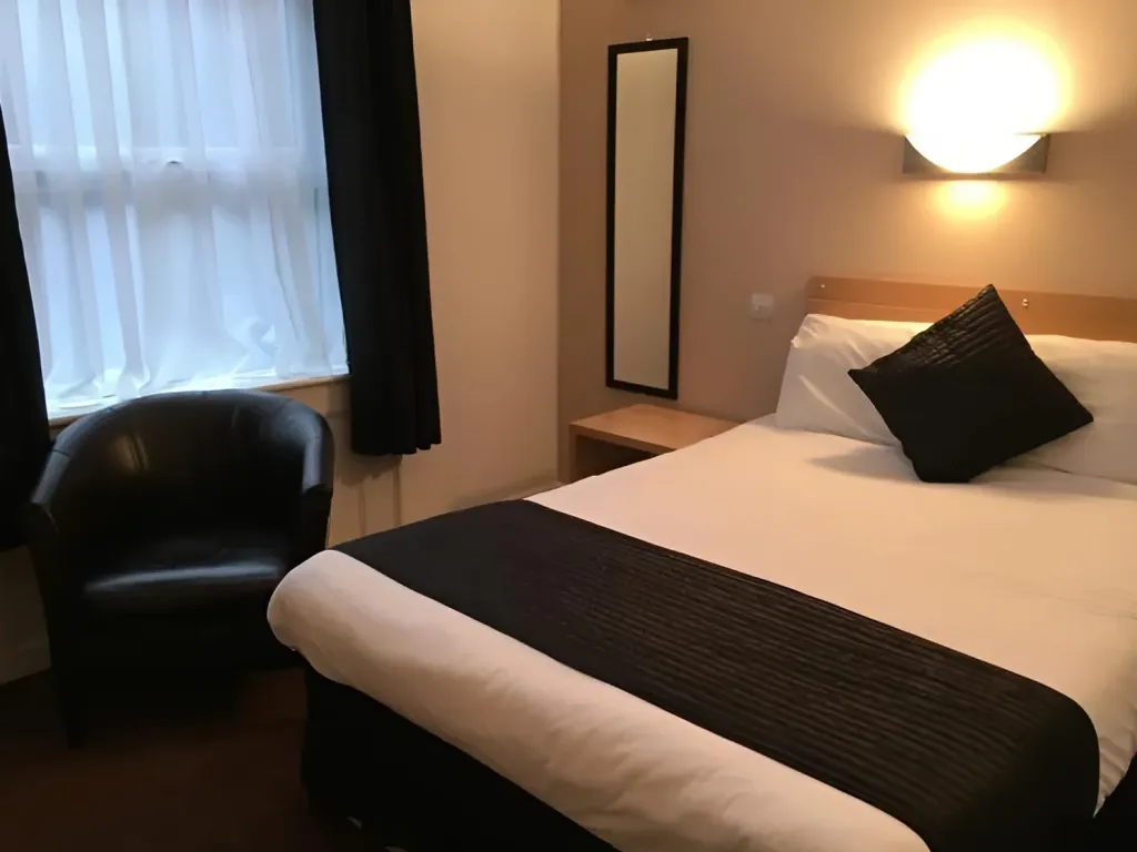 Hotel bedroom featuring a large bed with white linens, a dark accent pillow, a leather armchair, and a window with curtains
