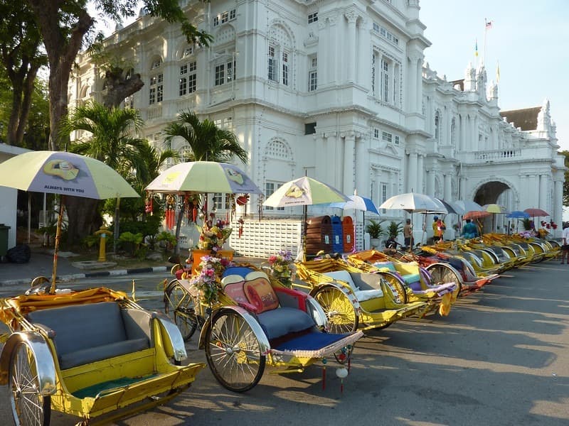 Rickshaws with umbrellas and flowers line a street before a white colonial building and palm trees.