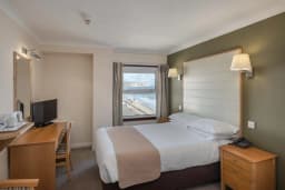 A hotel bedroom with a white bed, wooden furniture, and a window overlooking the ocean.