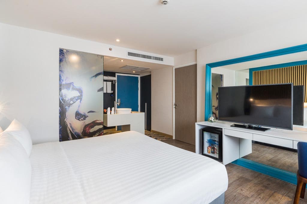 Hotel room with white bed, flat-screen TV, blue-framed mirror, and open bathroom area