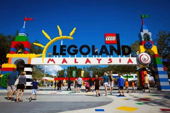 People enter a theme park under a large sign with block letters, a red LEGO logo, and colorful brick structures.