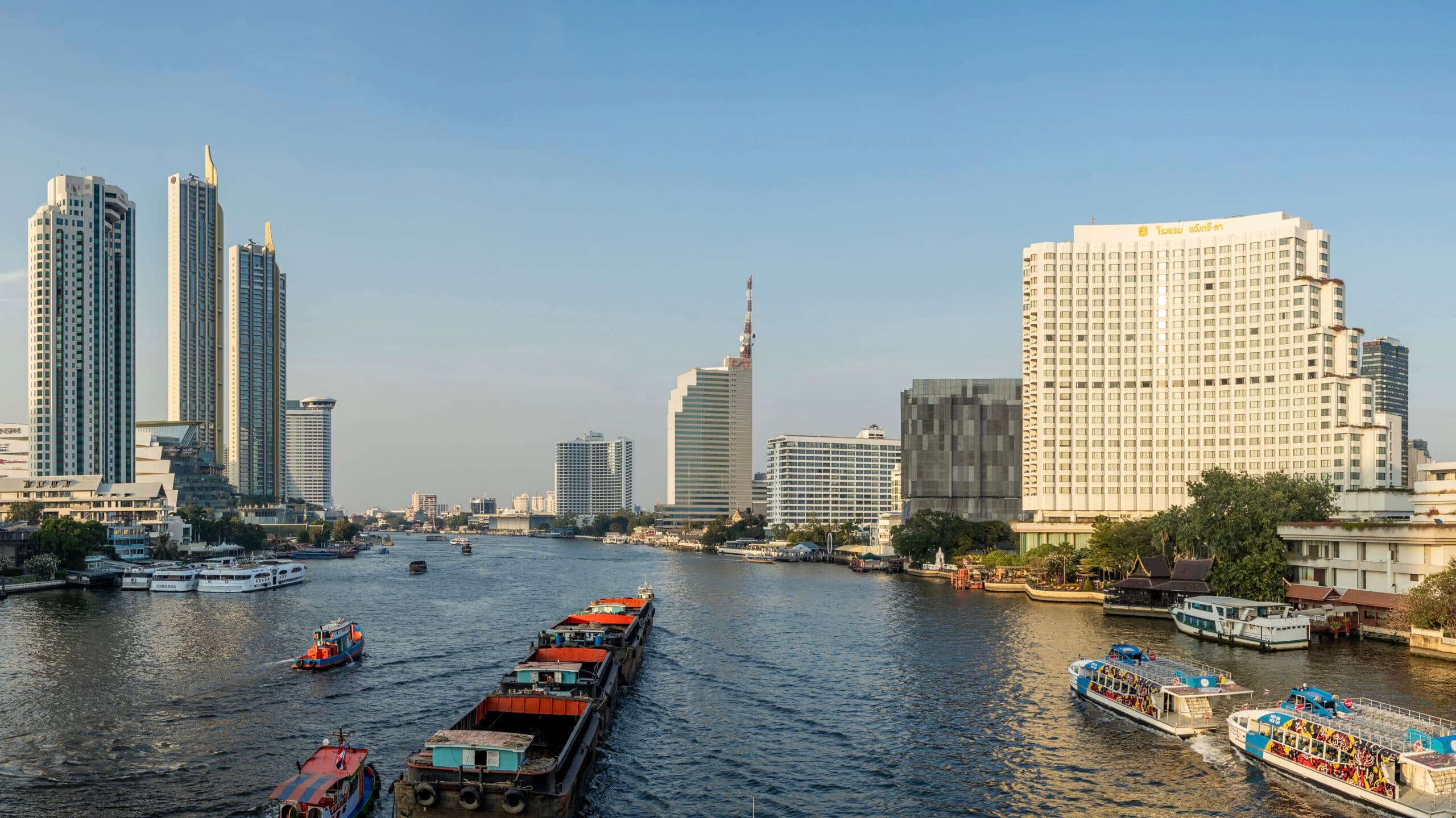 Boats and barges navigate a river lined with high-rise buildings and skyscrapers.
