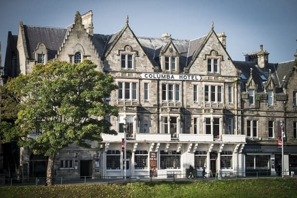 Stone building with "COLUMBA HOTEL" sign, a tree, and a grassy area in front.