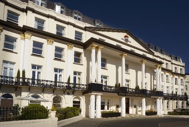 White hotel exterior with columns, balconies, and a crown emblem against a blue sky.