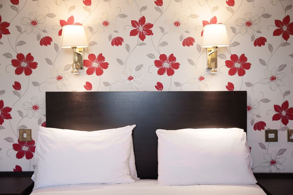 Bed with white pillows and dark headboard against floral wallpaper with two wall-mounted lamps