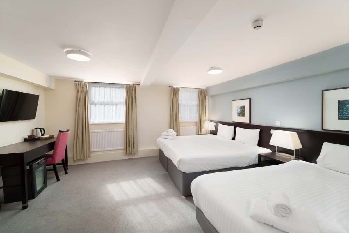 Hotel room with two double beds, a wall-mounted TV, desk, chair, and two curtained windows