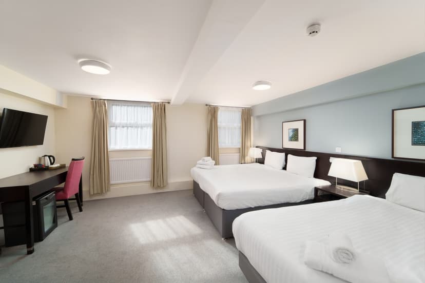 Hotel room with two double beds, a wall-mounted TV, desk, chair, and two curtained windows
