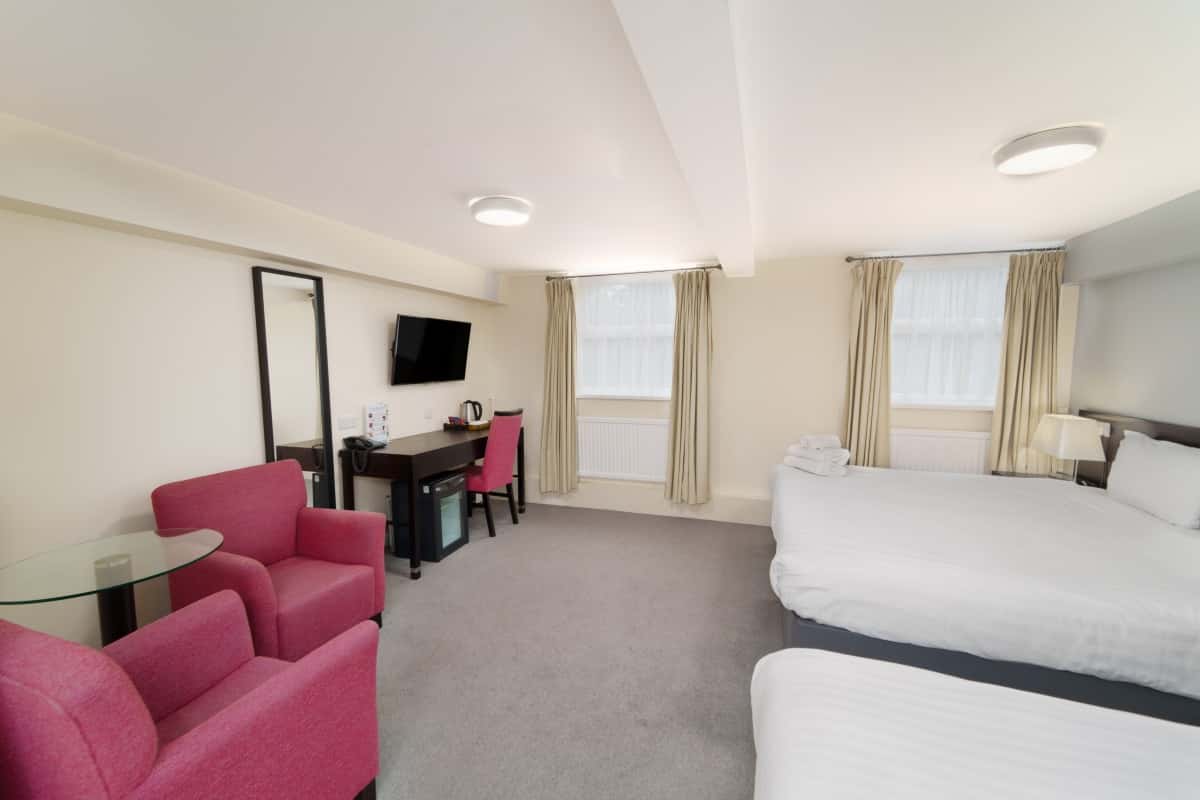 Hotel room with two beds, pink armchairs, a desk, mirror, and curtained windows