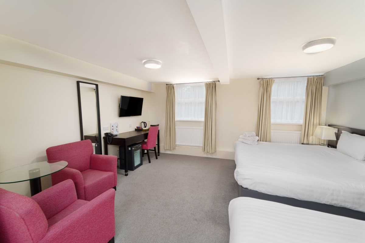 Hotel room with two beds, pink armchairs, a desk, mirror, and curtained windows