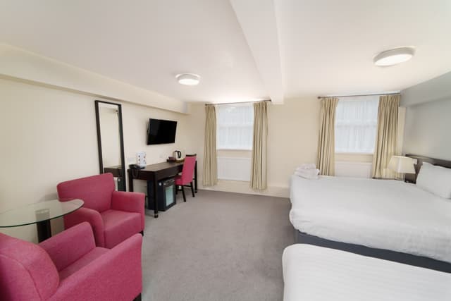 Hotel room with two beds, pink armchairs, a desk, mirror, and curtained windows