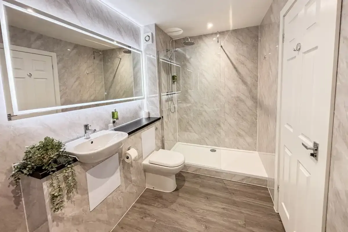 Bathroom with a walk-in shower, sink, toilet, and lit mirror on marble-tiled walls