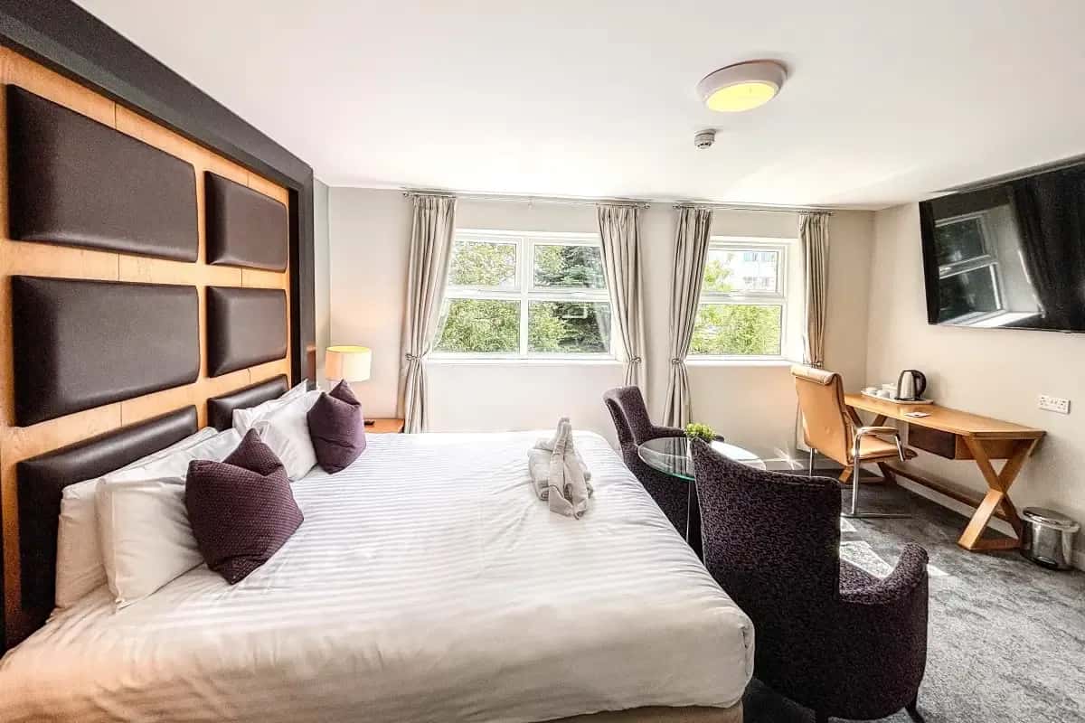 Hotel room with a padded headboard, double bed, seating area, desk, and wall-mounted TV