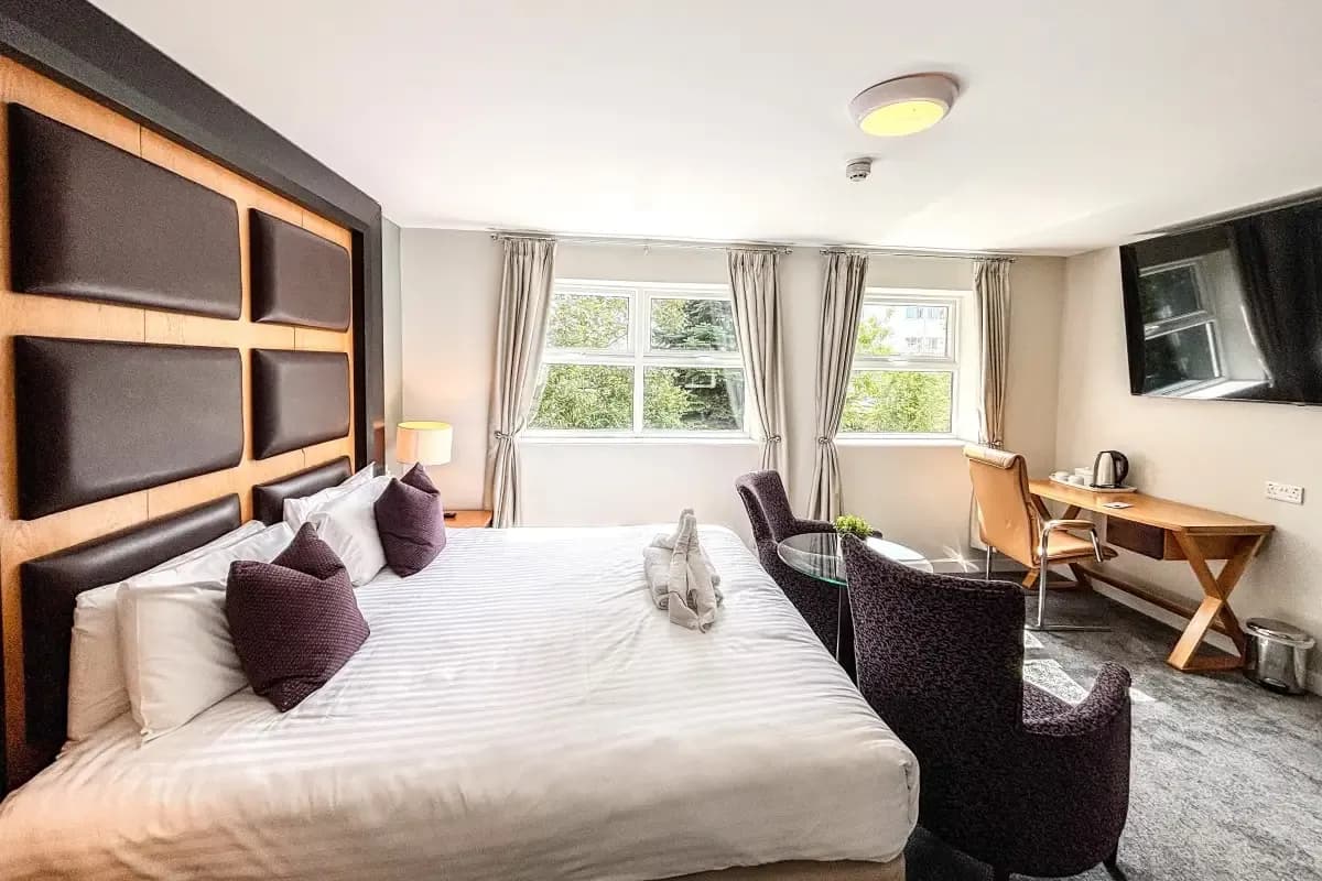Hotel room with a padded headboard, double bed, seating area, desk, and wall-mounted TV