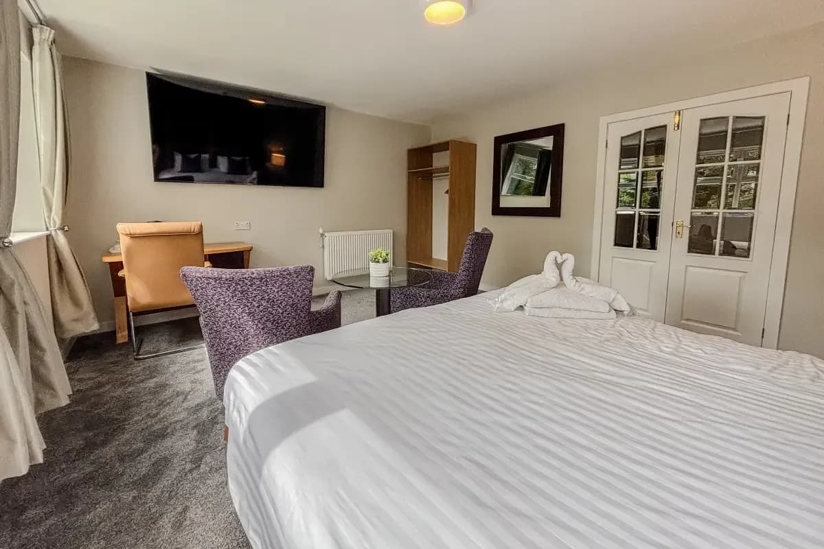 Hotel room with a white striped bed, folded towel swans, two armchairs, and a wall-mounted TV
