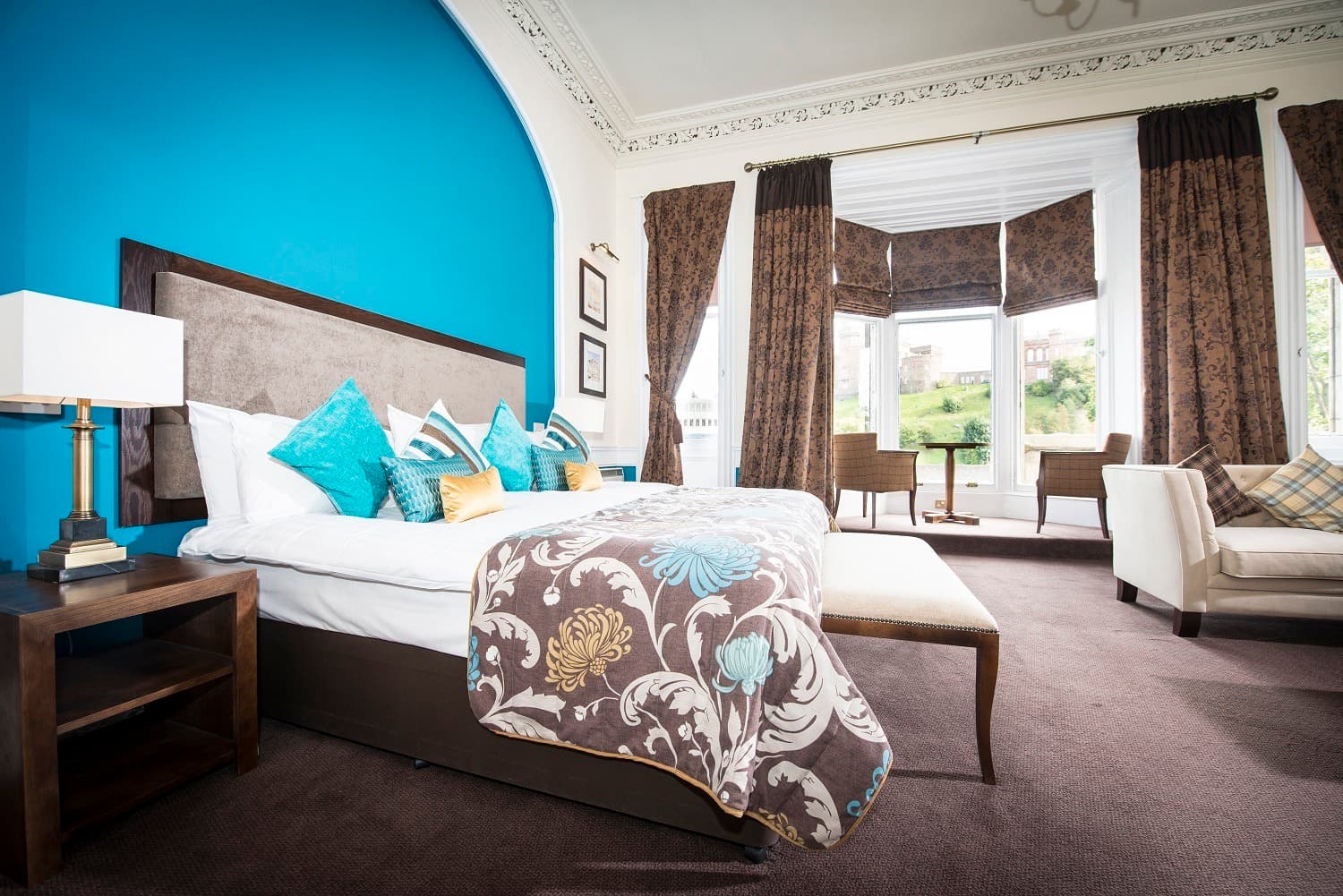 Hotel bedroom featuring a large bed with blue and patterned linens, a teal accent wall, and large windows with brown curtains