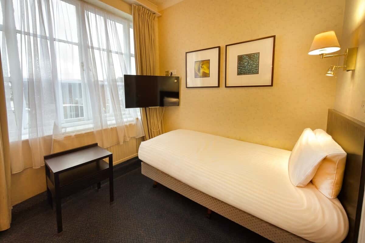 Single bed with white linens beside a wall-mounted TV, two framed pictures, and curtained windows