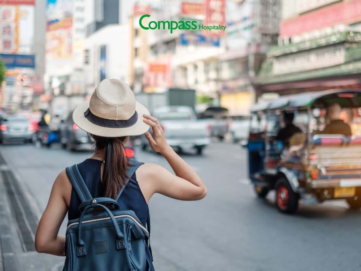 A woman wearing a backpack and straw hat stands on a city street with a tuk-tuk driving past.