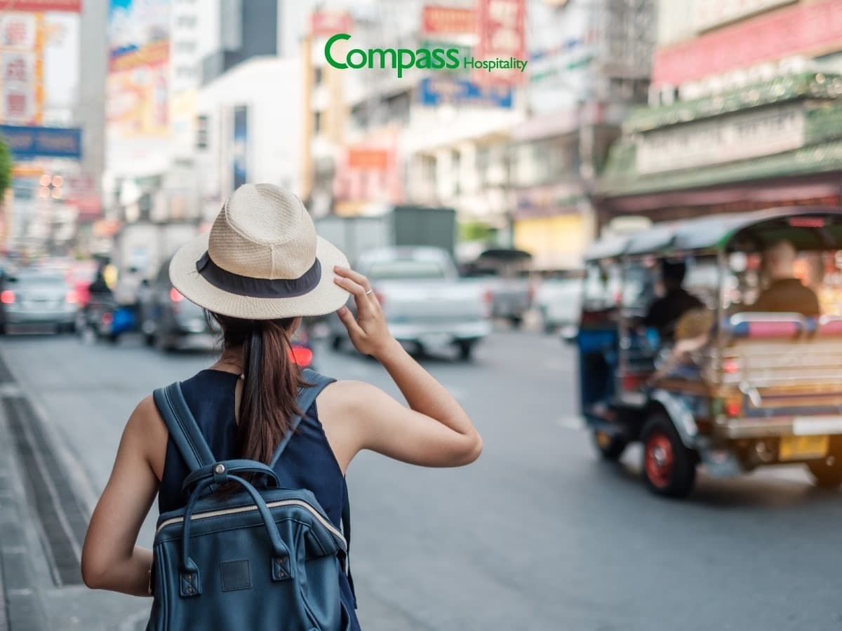 A woman wearing a backpack and straw hat stands on a city street with a tuk-tuk driving past.