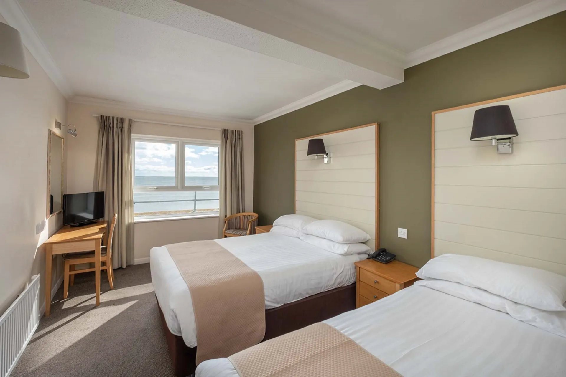 Two beds with white linens and tan runners sit in a room with a window overlooking the sea and a desk with a television