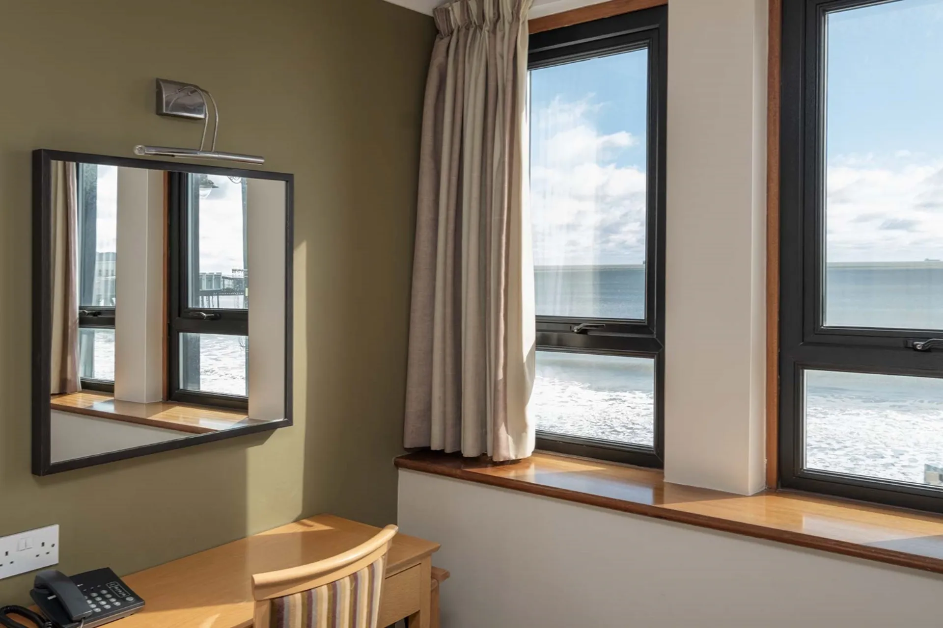 Window with ocean view and a mirror reflecting the view on an olive green wall next to a wooden desk