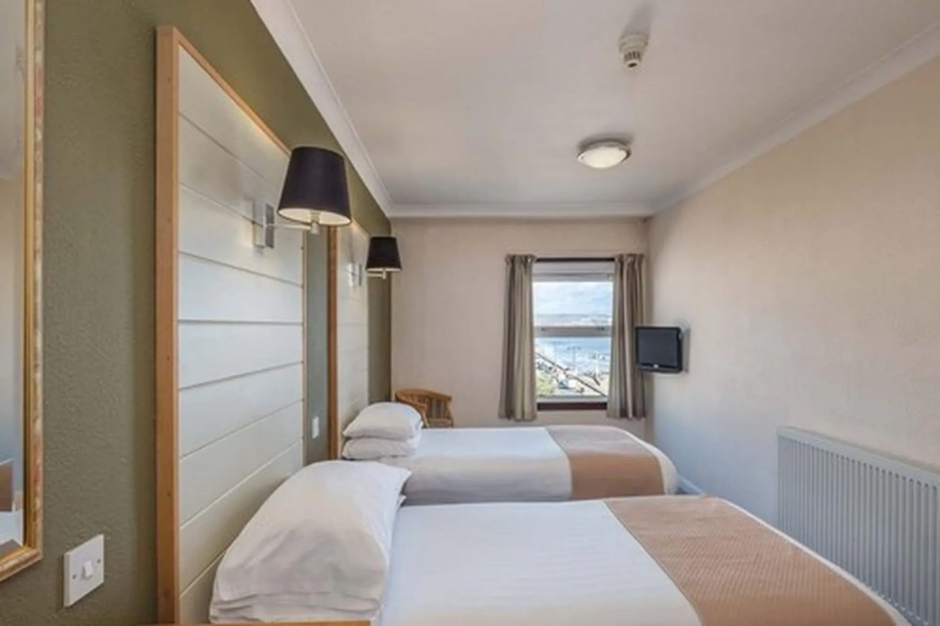 Two twin beds with white linens and tan runners sit in a room with a window and a television