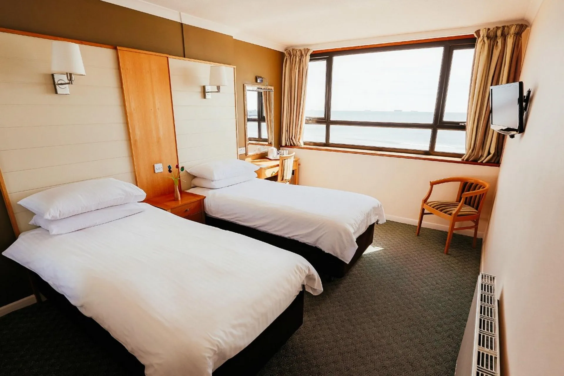 Two twin beds with white linens sit in a hotel room with a large window and a wooden chair