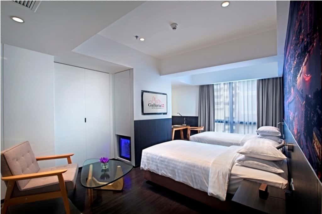 Twin beds with white linens beside a wardrobe, armchair, and wall-mounted TV showing "Galleria 12