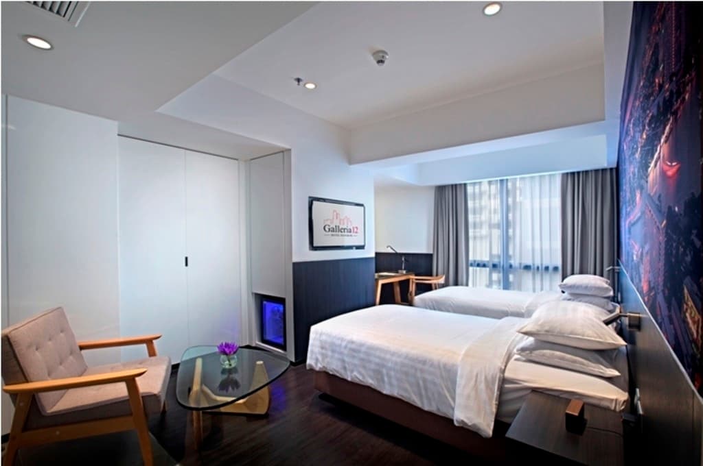 Twin beds with white linens beside a wardrobe, armchair, and wall-mounted TV showing "Galleria 12